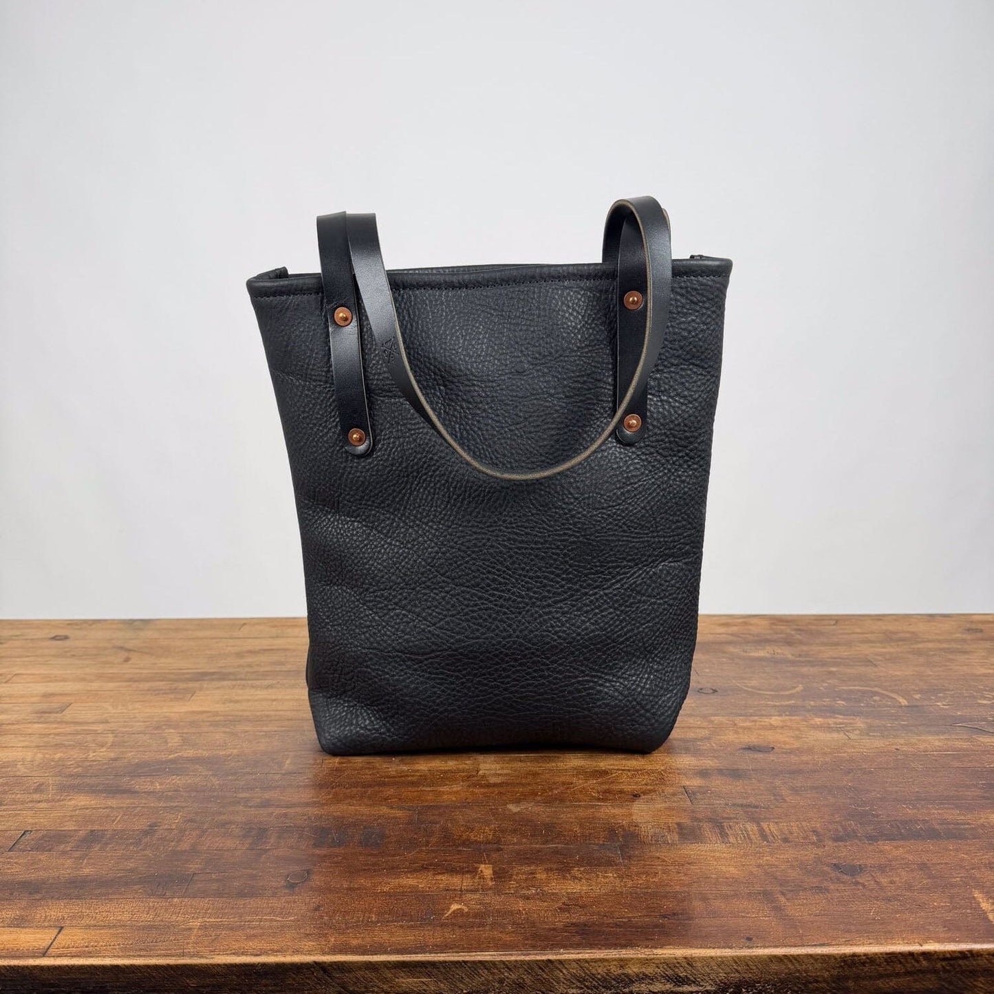 A black textured leather tote bag with dark brown leather handles and brass-tone rivets, standing on a rustic wooden table against a plain light gray wall.