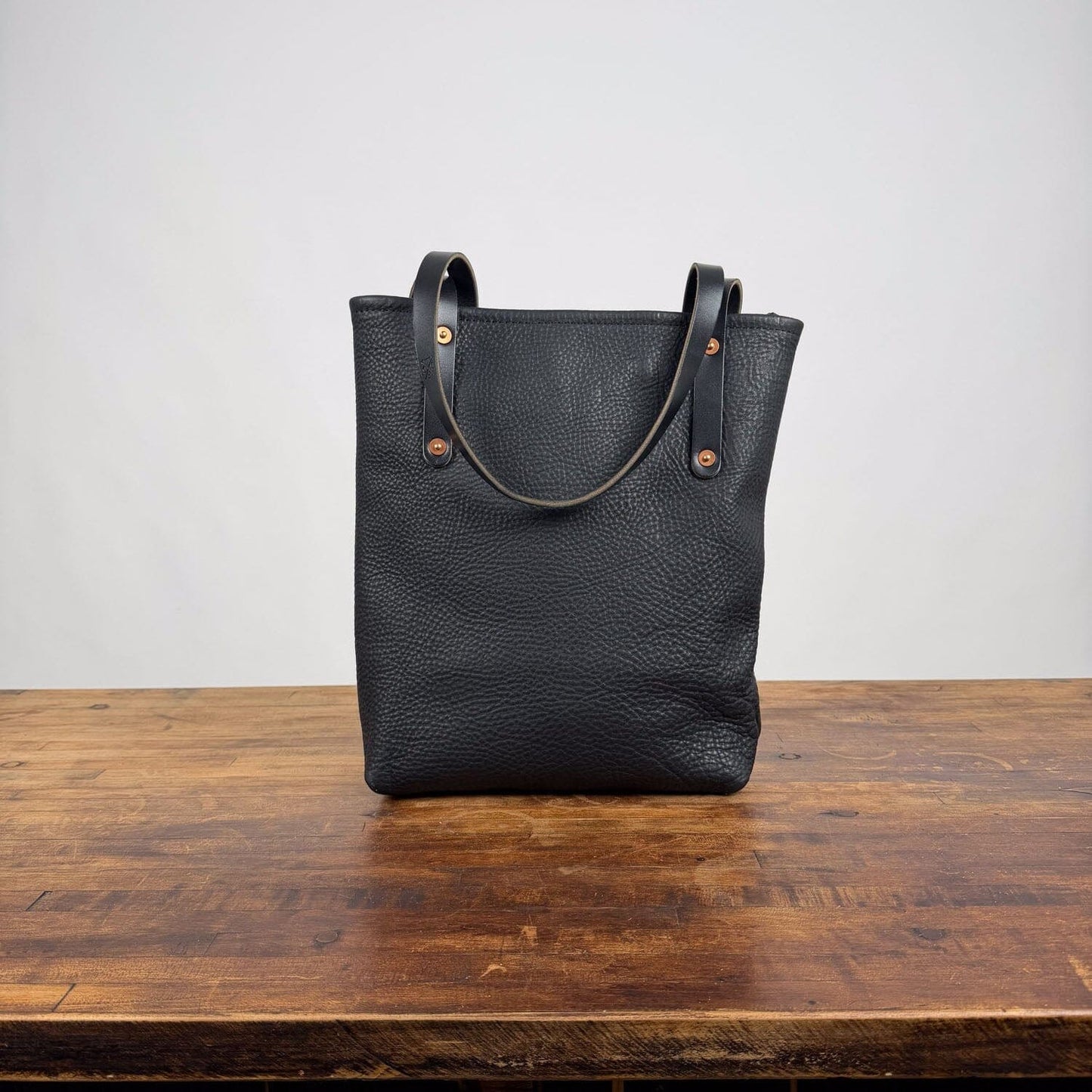 A black pebbled leather tote bag with double shoulder straps and brass-tone rivets, standing upright on a rustic wooden table against a plain light gray wall.