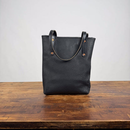 A black pebbled leather tote bag with double shoulder straps and brass-tone rivets, standing upright on a rustic wooden table against a plain light gray wall.
